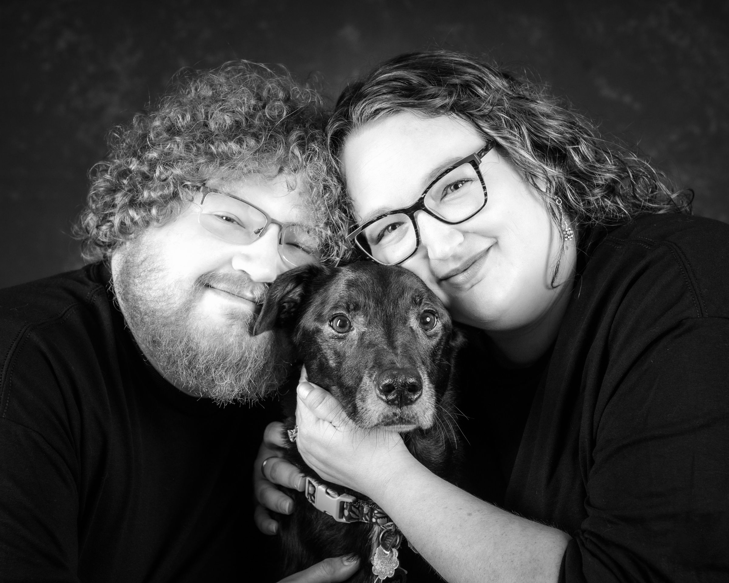 A couple pressing their cheeks to their grey-muzzled black dog