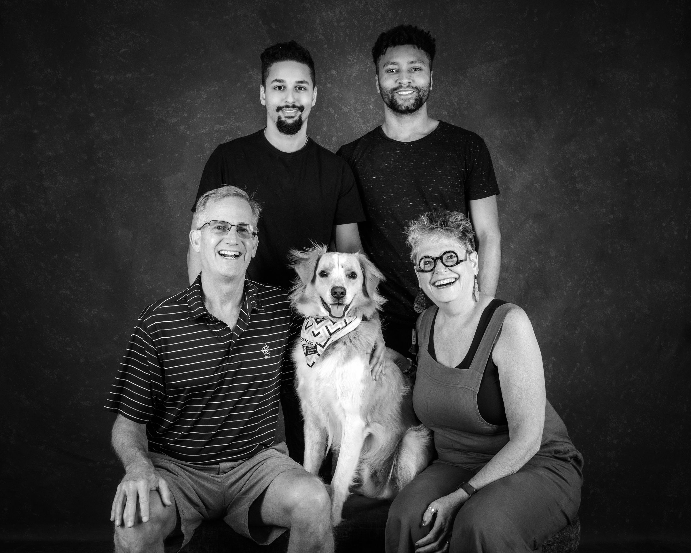 A family of four, two grown sons standing behind their parents, with their golden retriever in a bandana