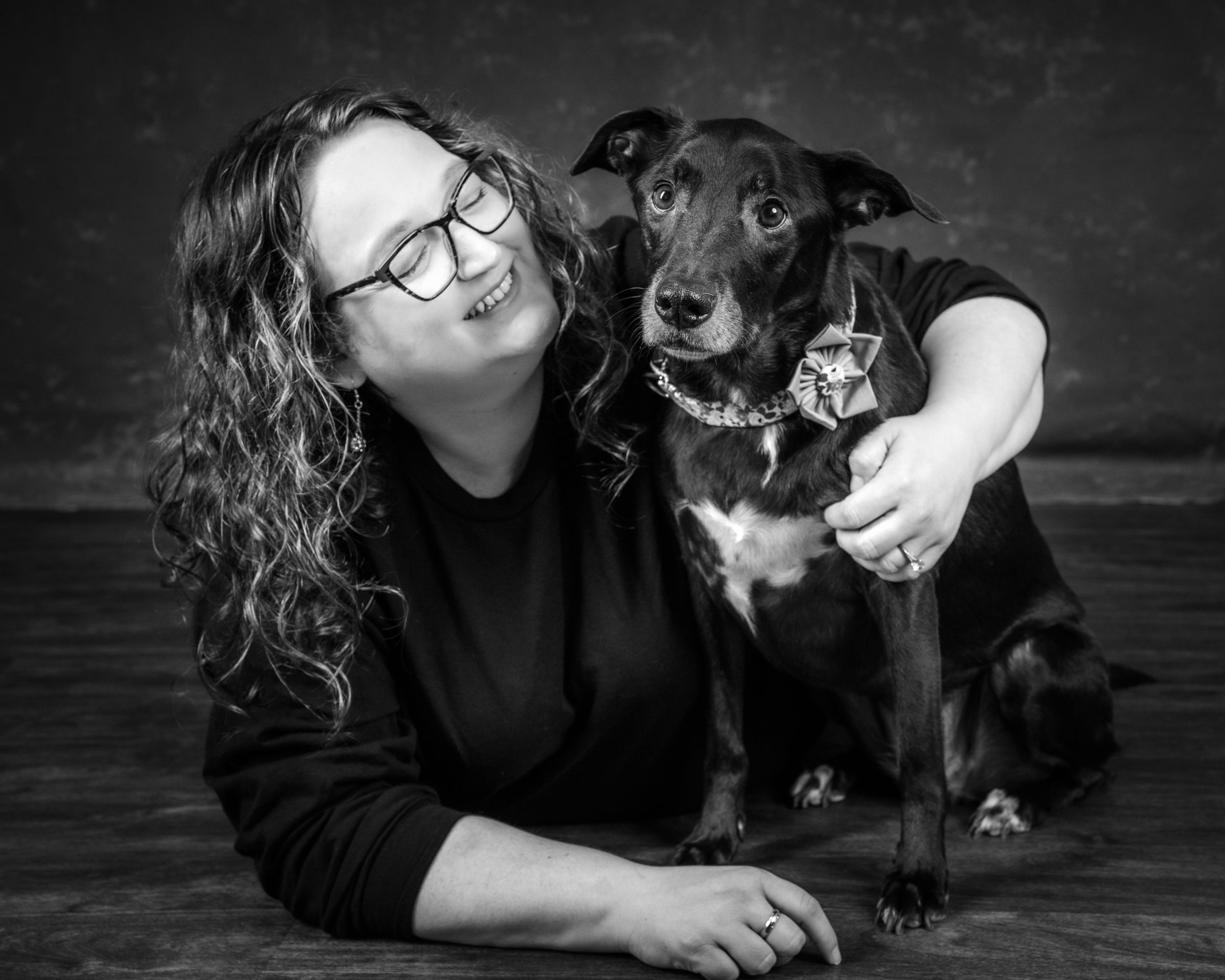 A woman laughing, her arm around her grey-muzzled black dog in a flower collar