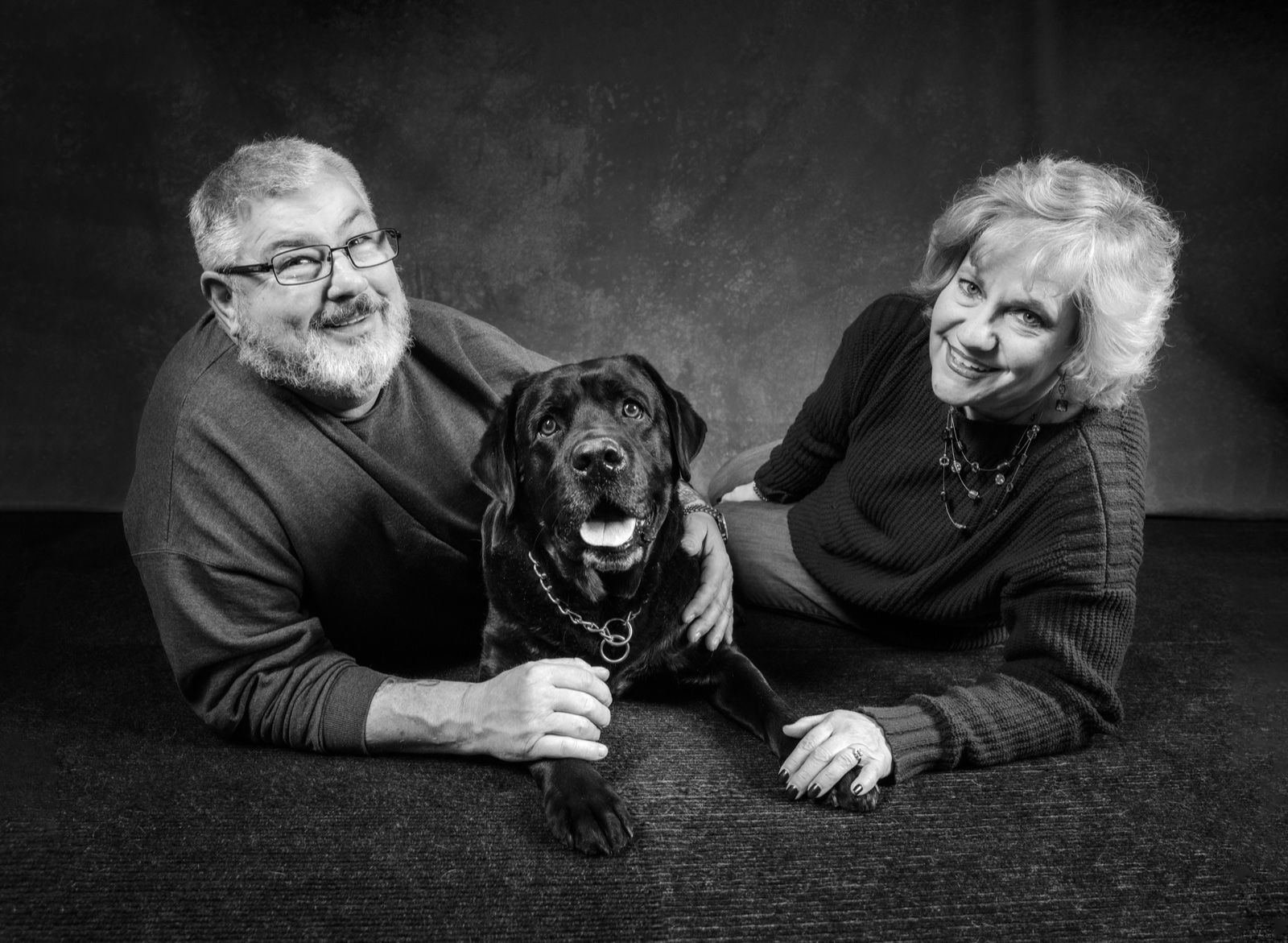 An older couple lying on the studio floor with their black lab between them
