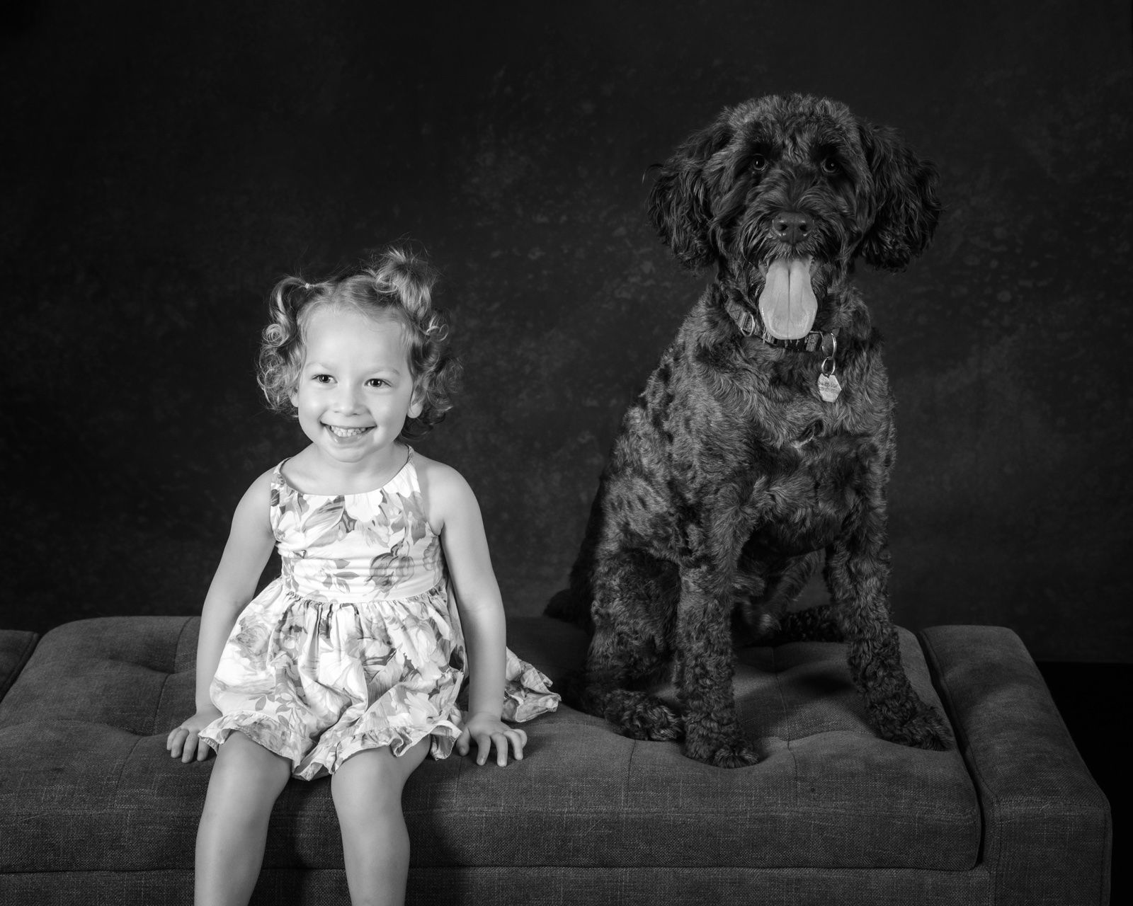 A toddler in a floral dress sitting beside a big black doodle