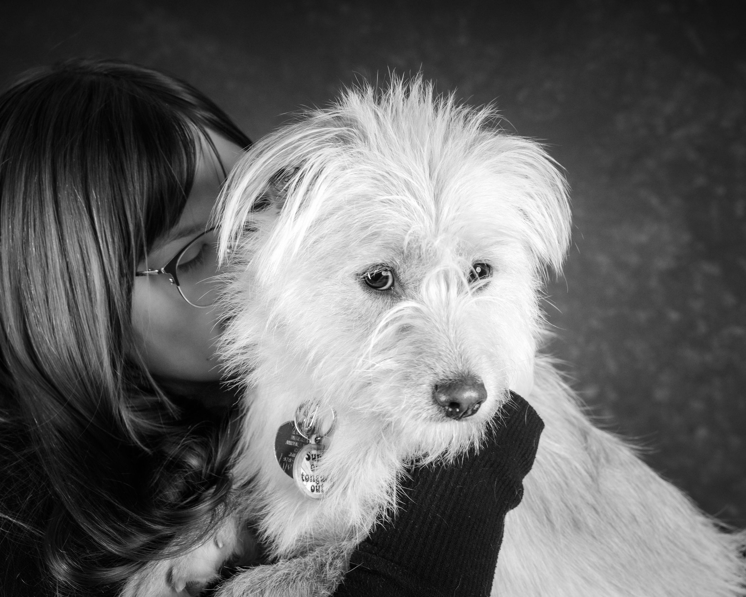 A woman kissing her scruffy white dog on the cheek while he looks at the camera