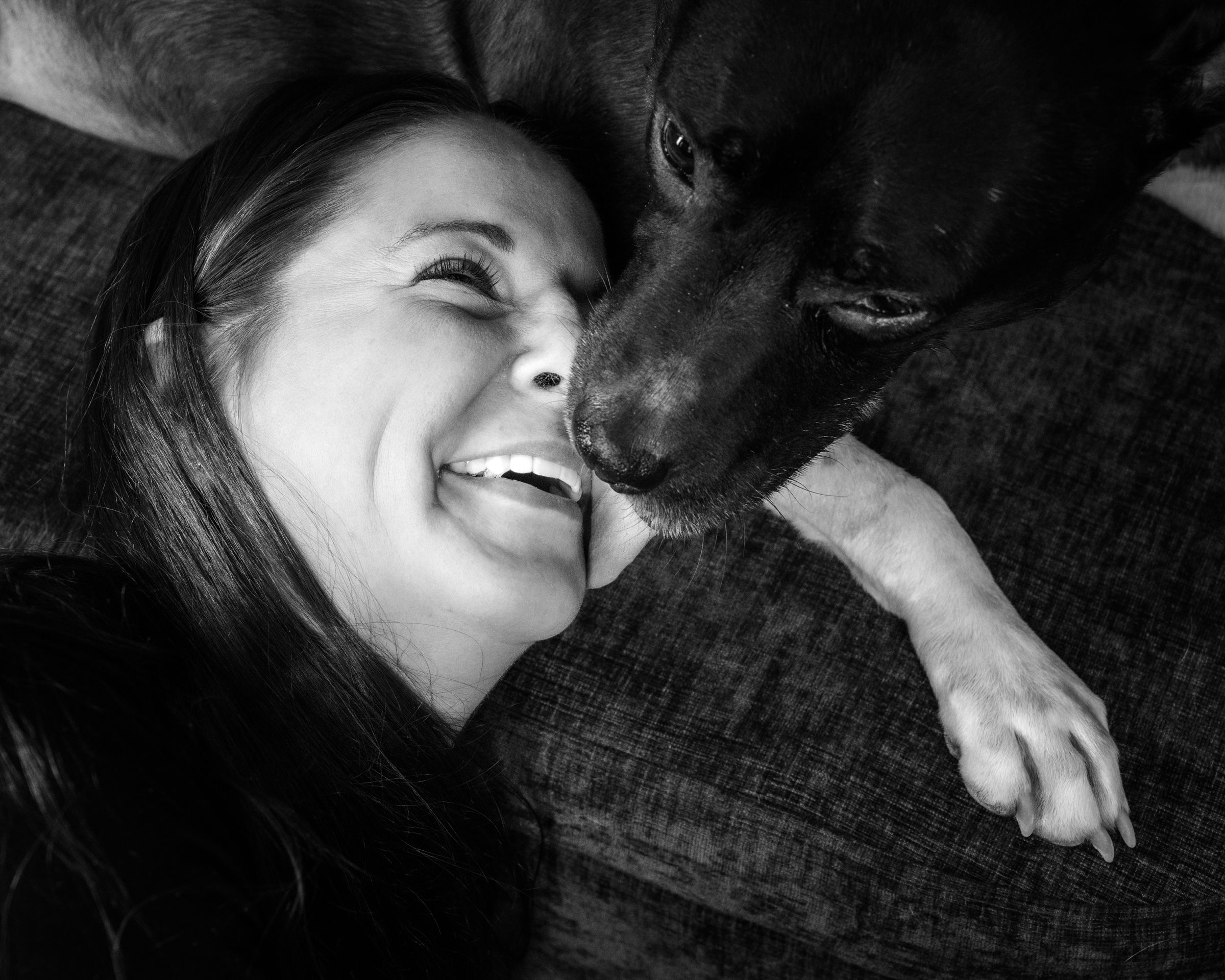A woman laughing with her eyes closed, her cheek pressed to her black dog's muzzle